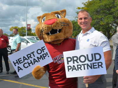 Mid-State Mascot Grit holding a sign that says "Heart of a Champ". Standing next to Grit is partner holding a sign that says "Proud Partner"