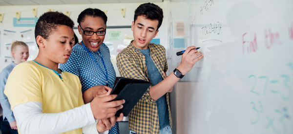 Two students and a teacher looking at a tablet. One student is also holding a marker up to a white board with math equations on it.