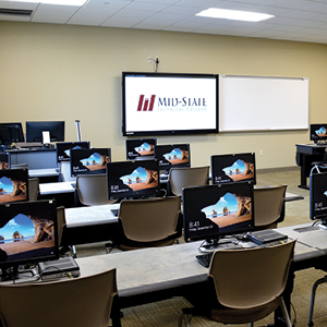 Computers on desks each with a chair, whiteboard and tv mounted in front of room