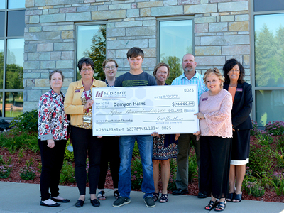 Damyon Hains holds up his $15,000 scholarship check with the assistance of Mid-State Foundation & Alumni Director Jill Steckbauer, left, and President Dr. Shelly Mondeik, right. Mid-State executive leadership team members also pictured, from left: Dr. Karen Brzezinski, vice president, Human Resources & Organizational Development; Dr. Mandy Lang, vice president, Student Services & Enrollment Management; Dr. Deb Stencil, vice president, Academics; Bradley Russell, vice president, Information Technology; and Dr. Bobbi Damrow, vice president, Workforce Development & Community Relations.