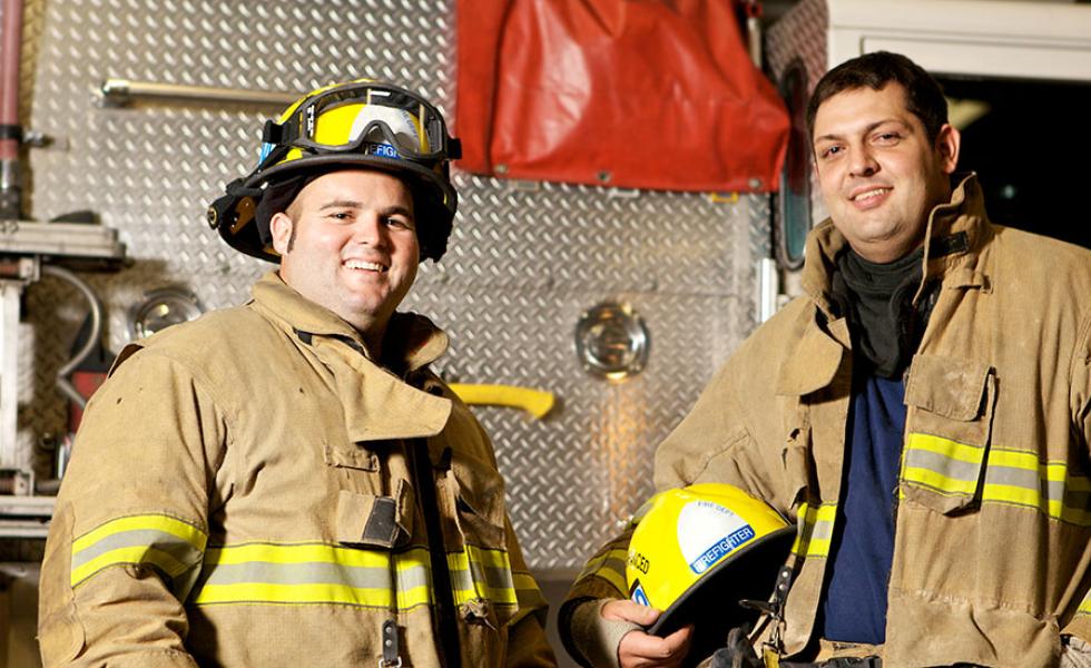 Two firefighters in full firefighting gear stand in front of a firetruck.