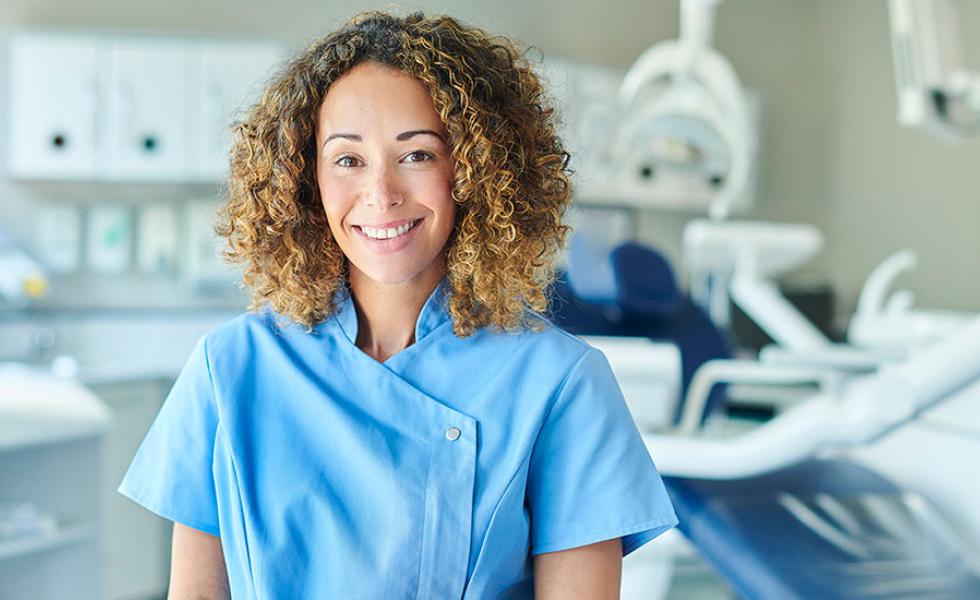 A woman wearing blue scrubs stands in front of a dental examination chair and equipment in a clinical setting.