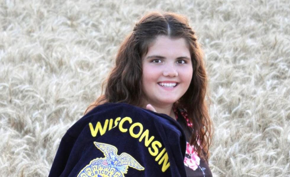 Macy Kloos in a field of wheat, holding a jacket with the word Wisconsin embroidered on the back