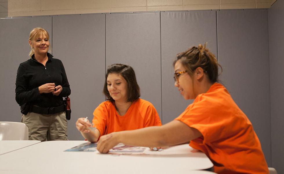 A jail officer stands nearby, watching two inmates work on a jigsaw puzzle at a table.