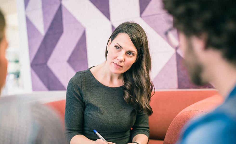 A counselor and clients sit across from each other on sofas. The counselor is listening and holding a pen.