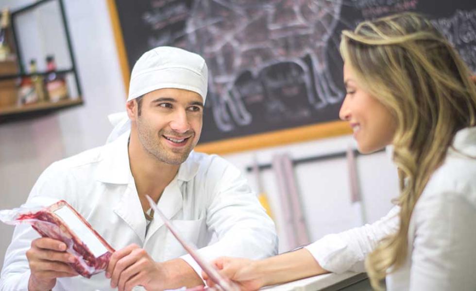 A man wearing a white smock and head covering holds a package of meat while leaning on a meat counter talking to a customer holding a piece of paper.