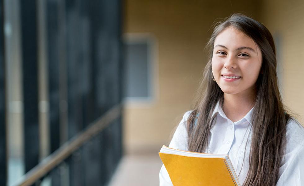 A young student stands in a hallway, wearing a backpack and clutching a spiral-bound notebook.