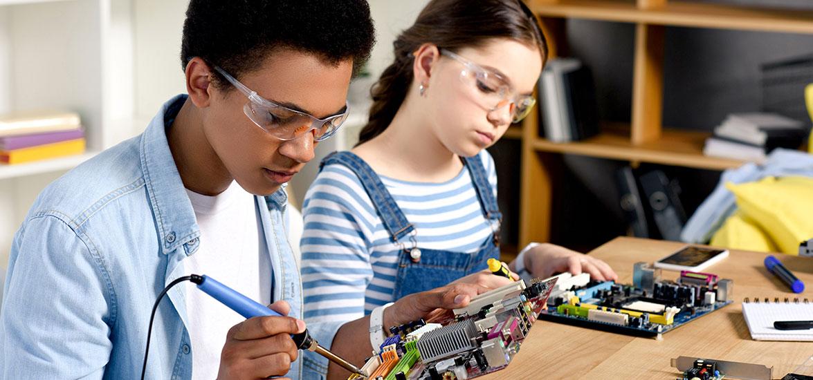 A boy and girl soldering digital circuit boards and wearing safety glasses.