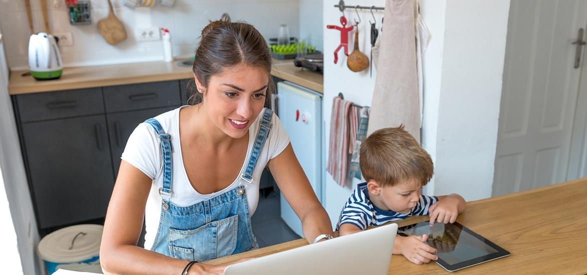 Woman sitting at a table looking at a laptop with a child sitting next to her looking at a tablet.