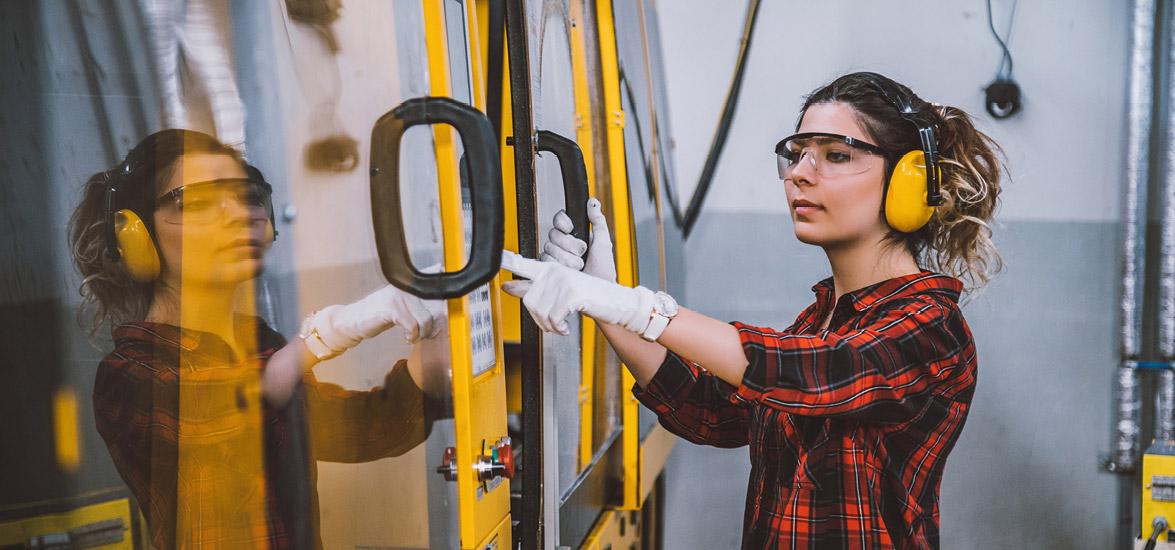 A female apprentice wearing hearing protection, goggles, and gloves operates a CNC machine