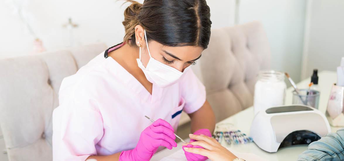 A masked nail technician wearing gloves performs a manicure on her client.