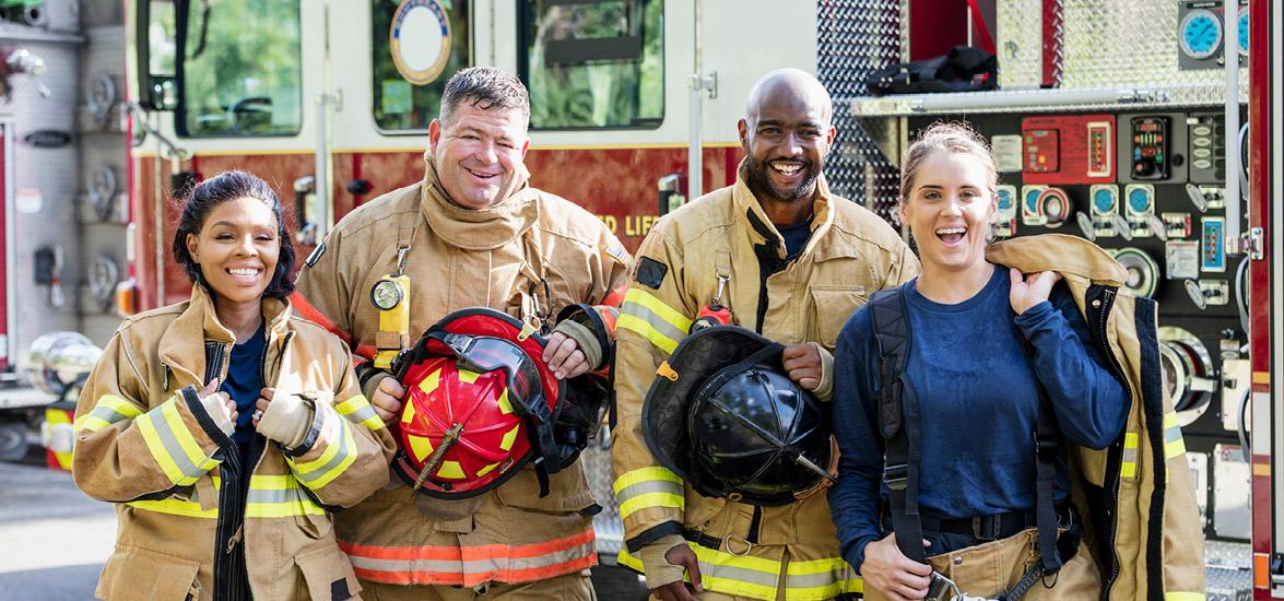 4 firefighters dressed up in full fire gear with their helmets off standing next to a firetruck smiling at the camera