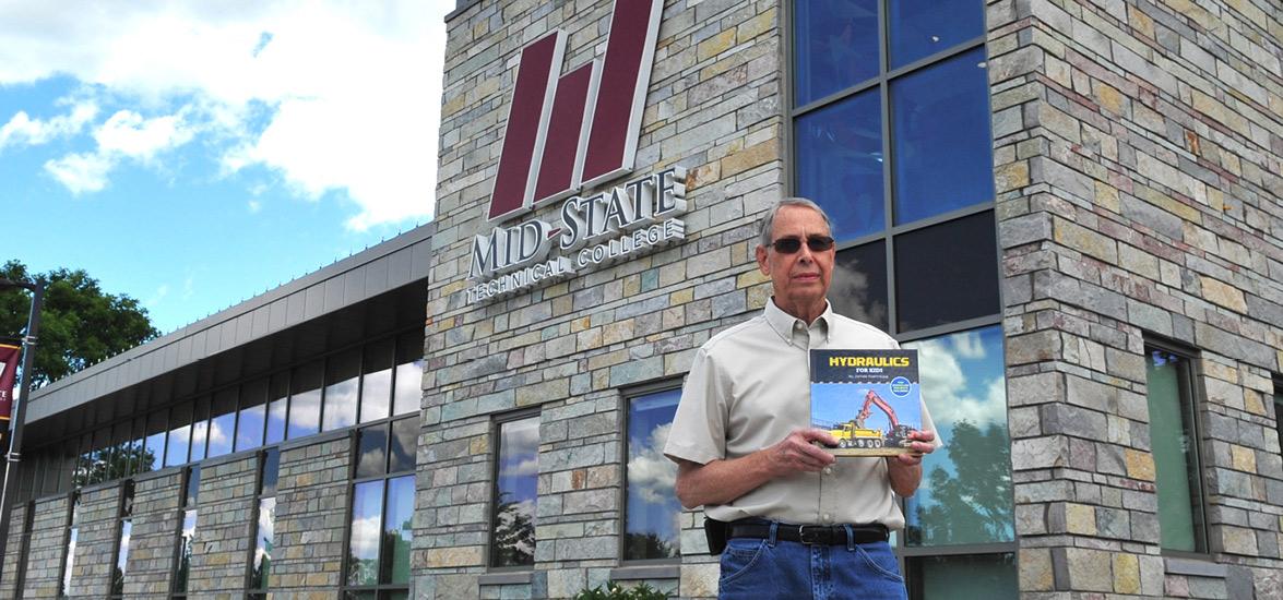 Retired Mid-State instructor Jim Koehntopp stands in front of the main entrance to Mid-State Technical College’s Wisconsin Rapids Campus with his recently published book, “Hydraulics for Kids.”