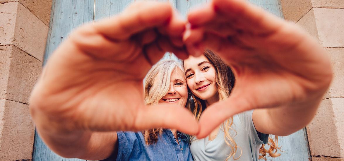 An older and younger woman stand arm-in-arm, forming a heart shape with their hands.