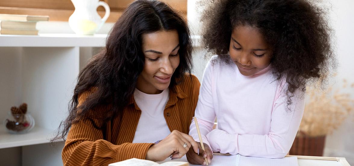 A woman helps a child write in a notebook.