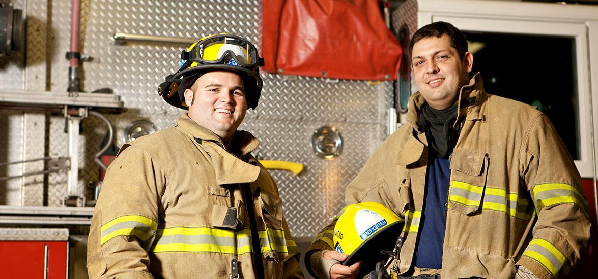 Two firefighters in full firefighting gear stand in front of a firetruck.