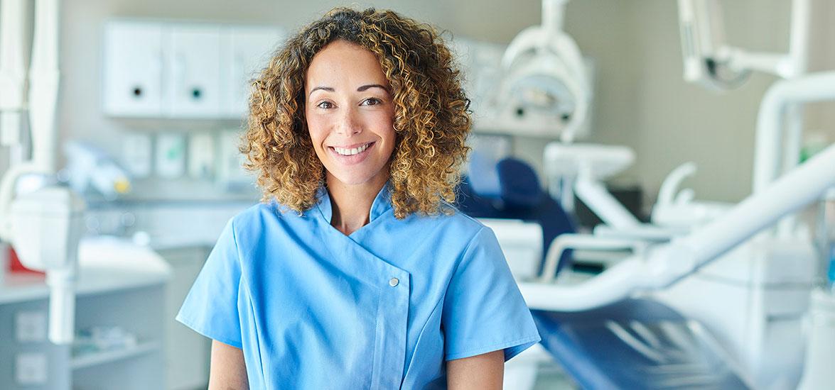 A woman wearing blue scrubs stands in front of a dental examination chair and equipment in a clinical setting.