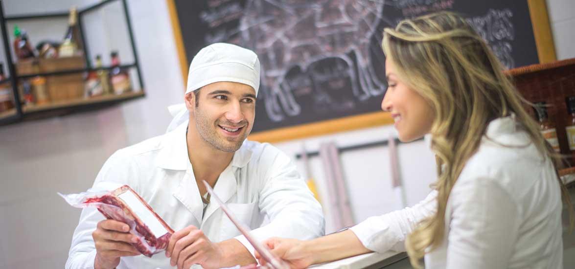 A man wearing a white smock and head covering holds a package of meat while leaning on a meat counter talking to a customer holding a piece of paper.