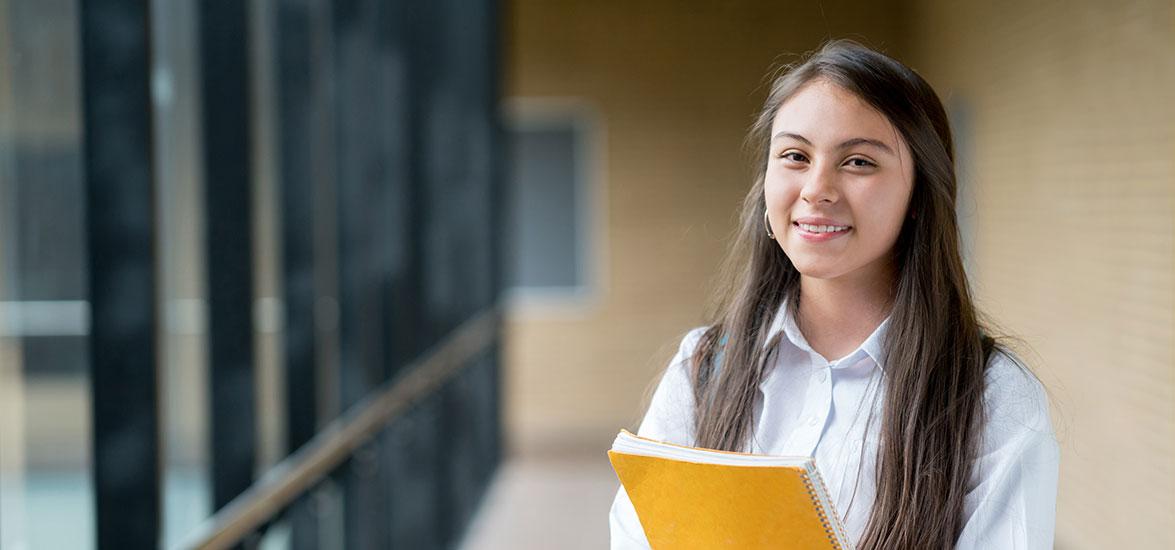 A young student stands in a hallway, wearing a backpack and clutching a spiral-bound notebook.