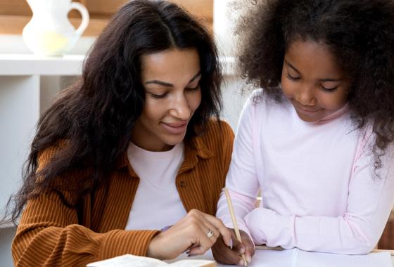 A woman helps a child write in a notebook.