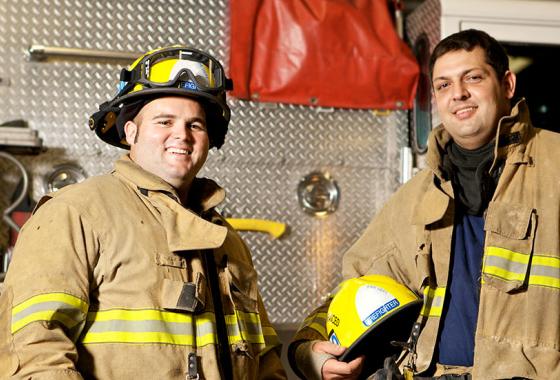 Two firefighters in full firefighting gear stand in front of a firetruck.