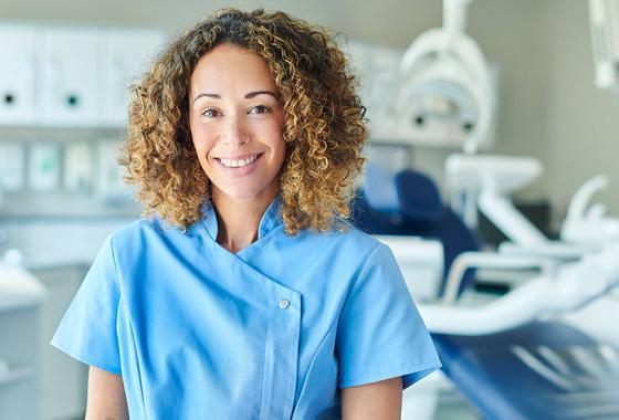 A woman wearing blue scrubs stands in front of a dental examination chair and equipment in a clinical setting.