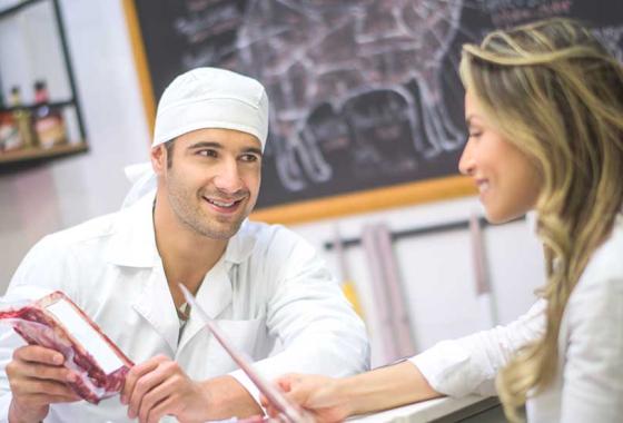 A man wearing a white smock and head covering holds a package of meat while leaning on a meat counter talking to a customer holding a piece of paper.