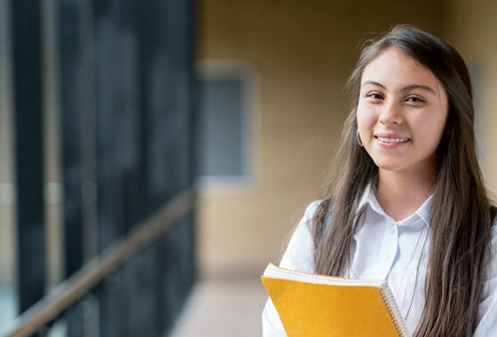 A young student stands in a hallway, wearing a backpack and clutching a spiral-bound notebook.