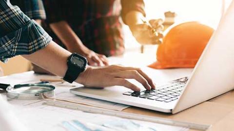 A person typing on a laptop sitting next to a hard hat.